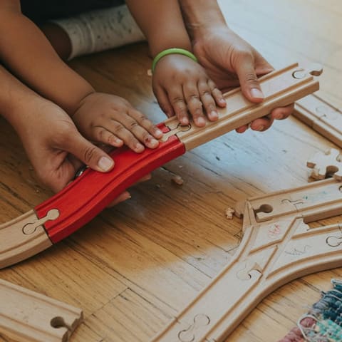 Close-up of a young child's hands and a parent's hands assembling a wooden train track on the floor